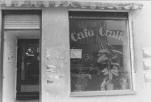s/w-Außenaufnahme eines Cafés. Im Schaufenster sind Grünpflanzen zu sehen, der Schriftzug am Fenster "Café Cralle" in Serifenbuchstaben mit dicken Rundungen an den Buchstabenenden.