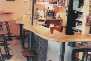 ein unregelmäßig geformter Holztresen, davor Barhocker, dahinter Geschirr, Kaffeemaschine.