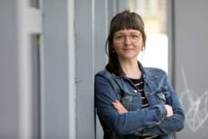 Frau mit Brille und Jeansjacke lehnt mit verschränkten Armen seitlich an Betonwand und schaut gerade in die Kamera. Heller Hintergrund.