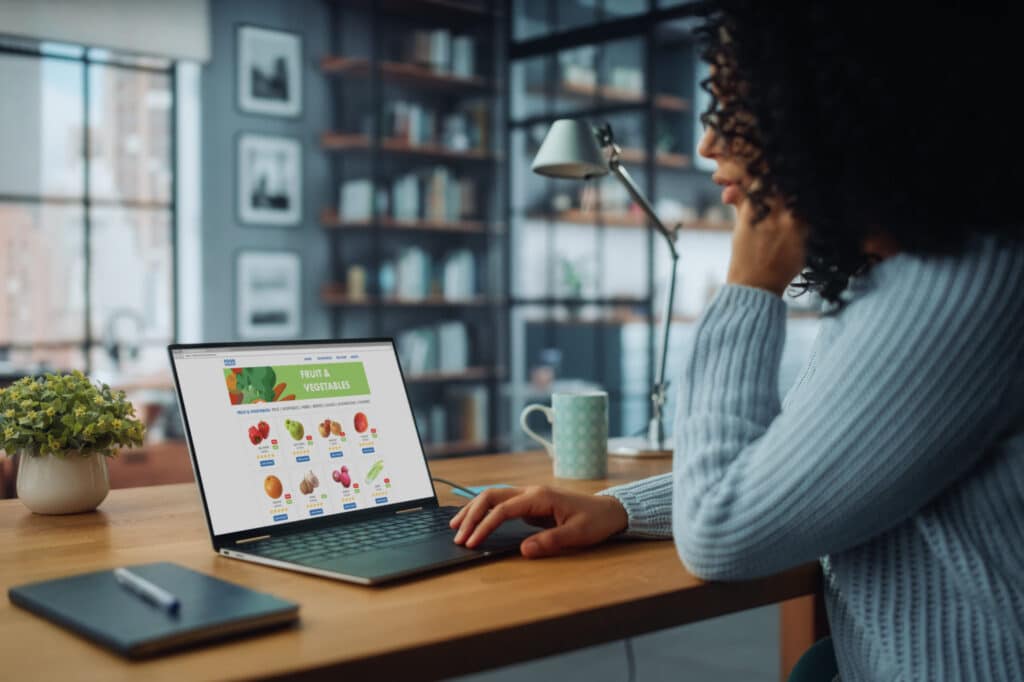 Back View Shot von einer jungen Frau mit Laptop-Computer und Bestellung von frischem Gemüse durch Lebensmittel Lieferung App Web-Seite. Daytime in Bright Studio Apartment mit großen Fenstern.